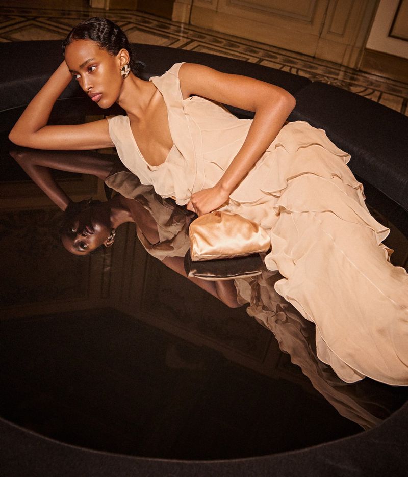 Elegant woman in a flowing beige dress reclines near reflective surface with a small clutch, illuminated by soft, ambient lighting.