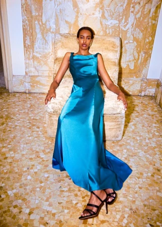 Model wearing a long teal satin gown seated in an upholstered armchair against an ornate interior wall.
