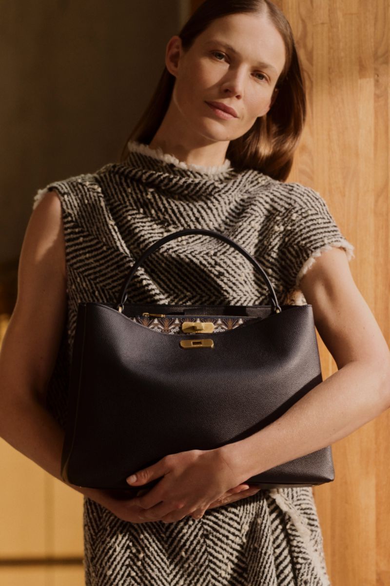 Elegant woman showcasing a black leather handbag with a golden clasp, dressed in a chic patterned dress against a warm wood backdrop.