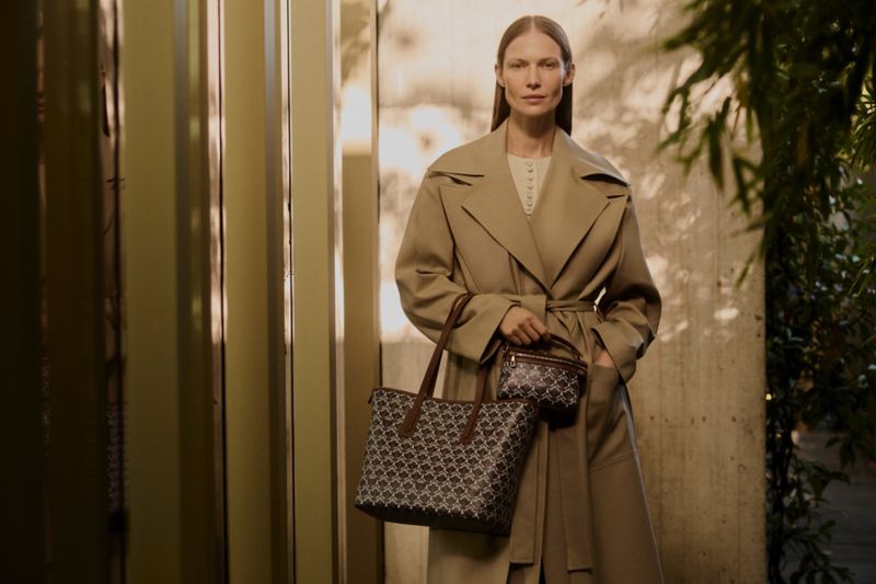 A stylish woman in a tan trench coat poses with a patterned tote bag and small purse, complemented by a muted beige background with greenery.