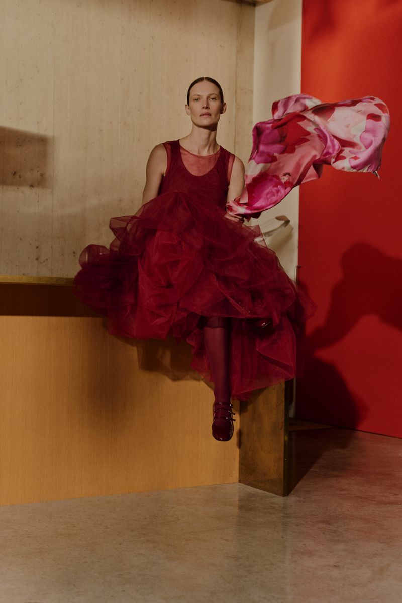 A slender woman sits on a ledge in a red tulle dress with a flowing floral scarf, with an indoor studio environment with orange and red walls.