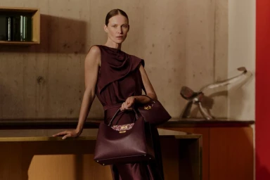 Model in dark burgundy dress leaning on a wooden counter holding matching handbags against a concrete wall