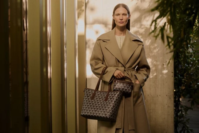 Model standing outdoors wearing a camel trench coat holding patterned handbags for Liberty AW24 campaign