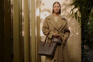 Model standing outdoors wearing a camel trench coat holding patterned handbags for Liberty AW24 campaign