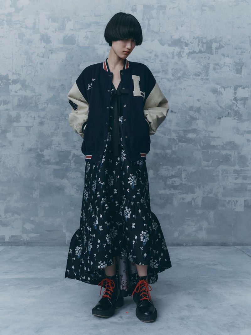 A woman models a varsity jacket and floral dress in a studio, her hands in her pockets and her gaze directed slightly downward.