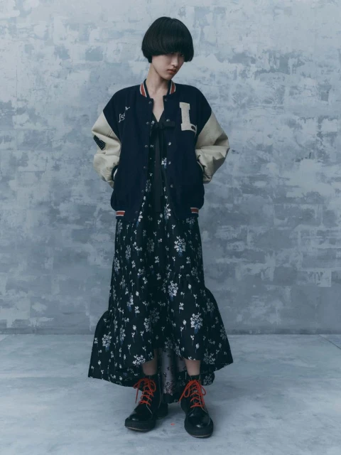 A woman models a varsity jacket and floral dress in a studio, her hands in her pockets and her gaze directed slightly downward.