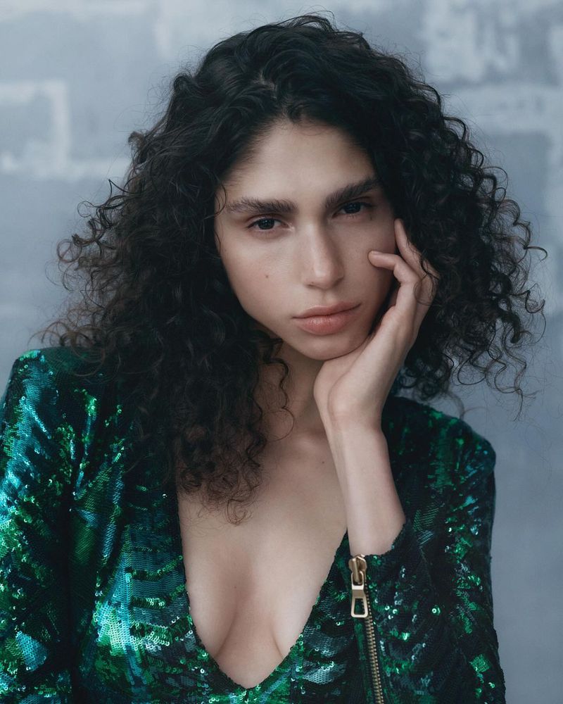 Portrait of a woman with curly hair wearing a green sequin blazer, hand on face in a studio setting with textured grey background.
