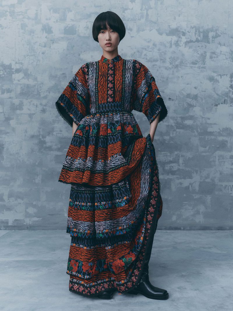 Model wearing a patterned tiered dress with rich orange and blue stripes against a textured grey background
