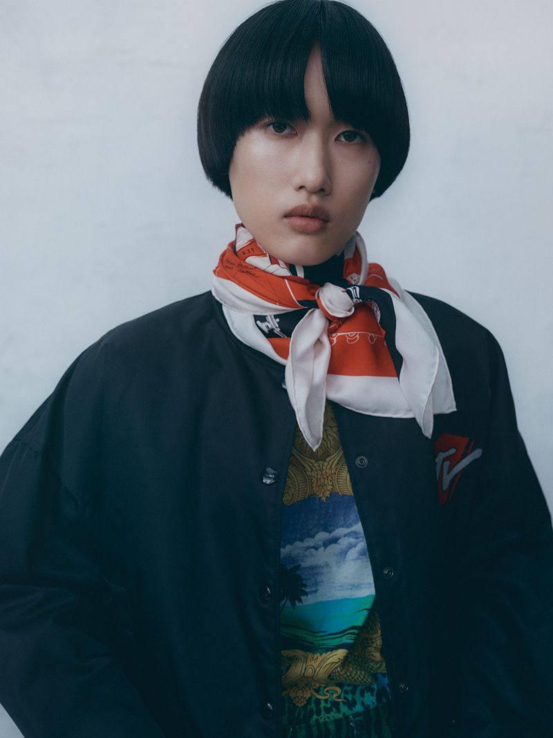 Close-up of a young adult woman with a bowl haircut, wearing a black jacket and a patterned silk scarf against a neutral backdrop.