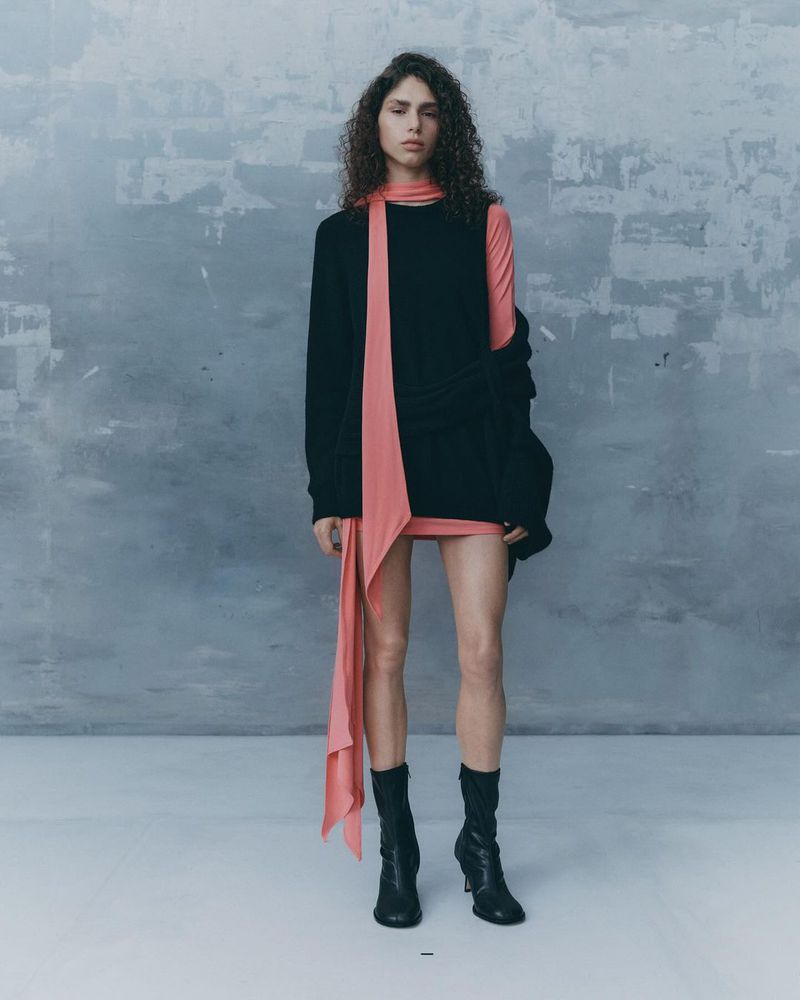 Model with curly dark hair wearing a black sleeveless top, pink scarf, and black boots against a textured grey wall