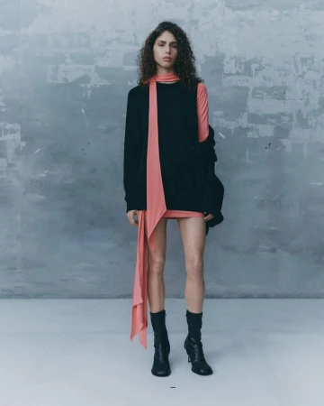 Model with curly dark hair wearing a black sleeveless top, pink scarf, and black boots against a textured grey wall