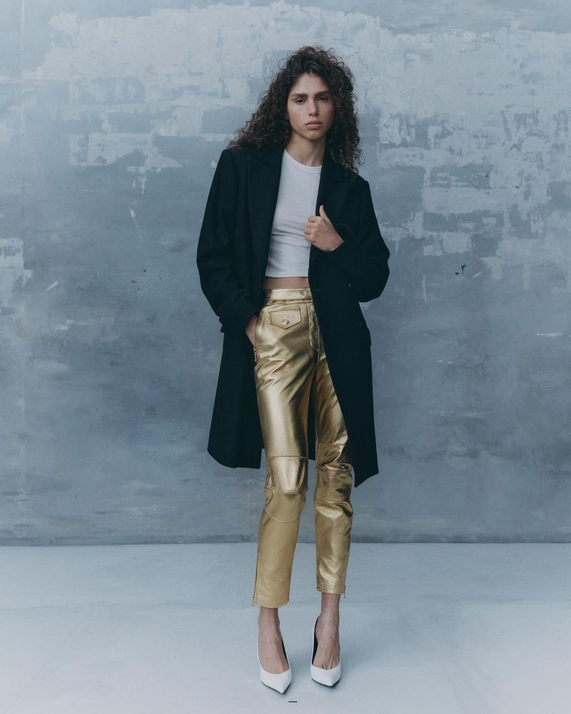 Full-length portrait of a curly-haired woman in a black coat, white top, gold pants, and white heels, against a textured grey wall.
