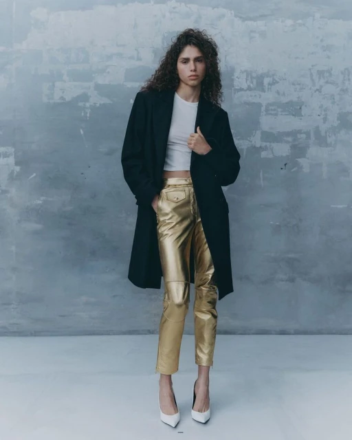 Full-length portrait of a curly-haired woman in a black coat, white top, gold pants, and white heels, against a textured grey wall.