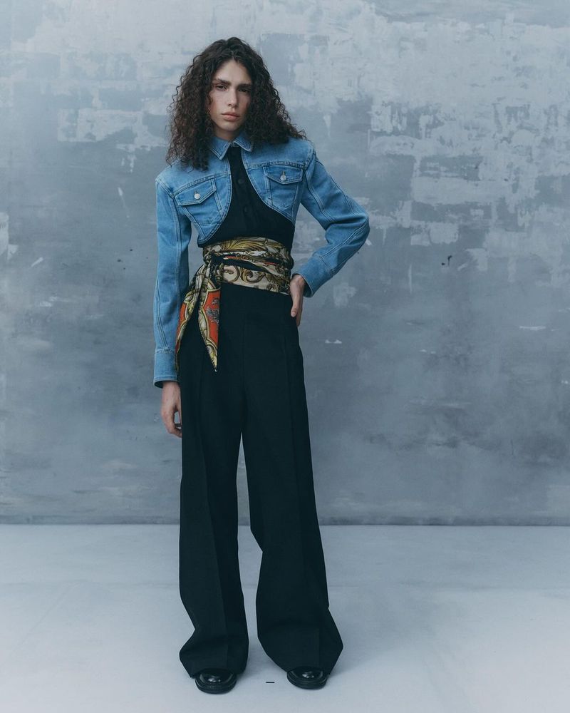 Model with long curly hair wearing a cropped denim jacket over a black jumpsuit and patterned scarf