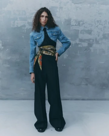 Model with long curly hair wearing a cropped denim jacket over a black jumpsuit and patterned scarf