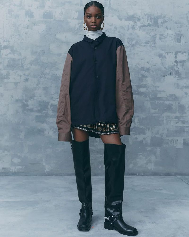 Fashion portrait of a Black female adult wearing a color-blocked shirt, skirt, knee-high boots, and hoop earrings against a textured background.