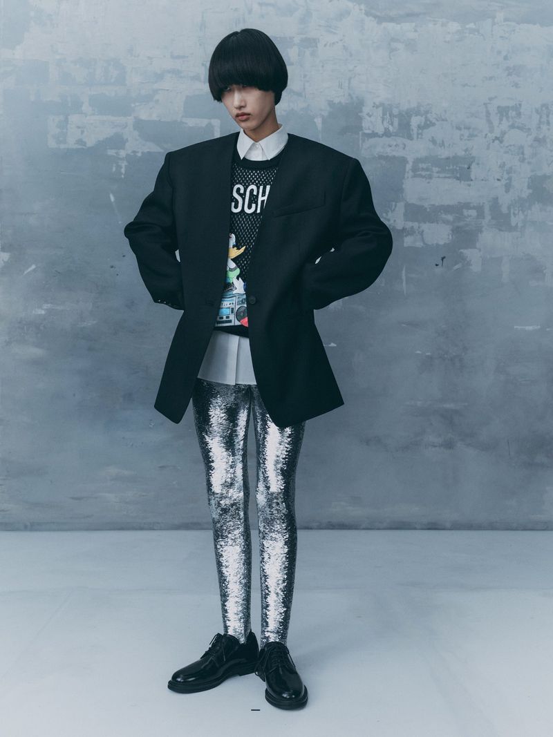 A person in a black blazer, patterned top, and silver leggings stands in front of a grey textured wall, illuminated by studio lighting.