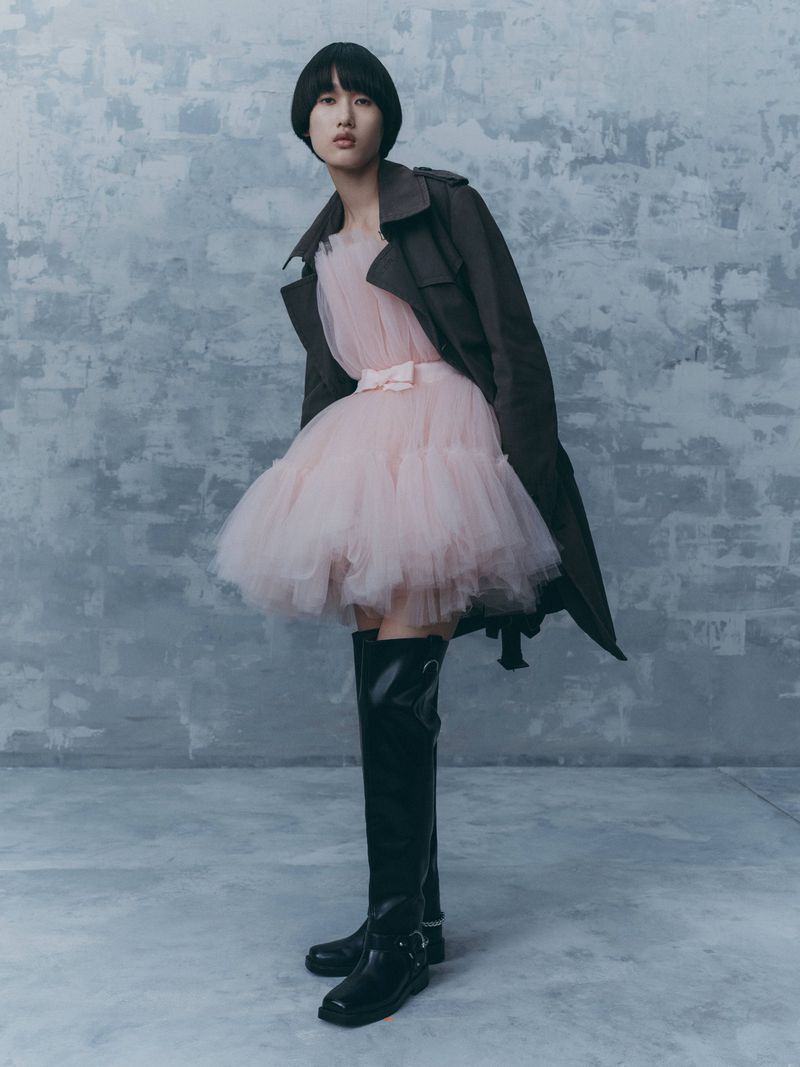 A stylish adult female fashion model poses in a short pink dress, trench coat, and thigh-high boots against a textured grey backdrop.