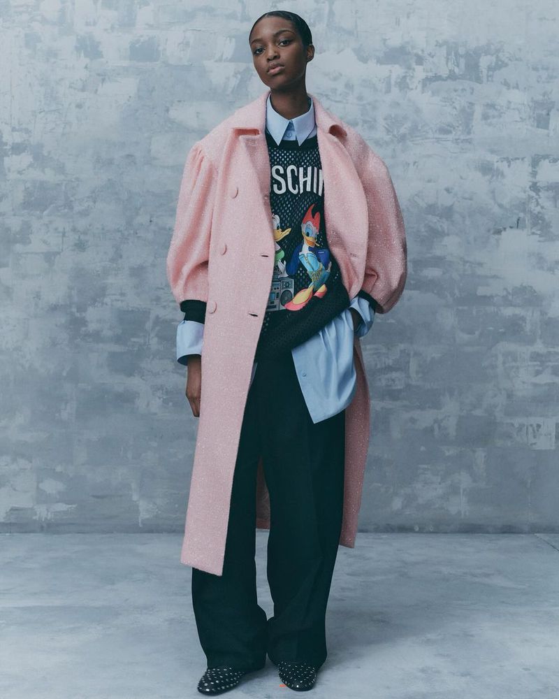 Editorial fashion shot of a woman in a light pink coat and black slacks standing in front of a gray textured wall, looking at the camera.