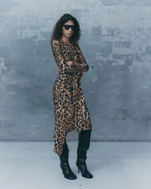 Woman with curly hair wearing sunglasses and a leopard print dress, standing indoors against a textured grey wall in a fashion pose.