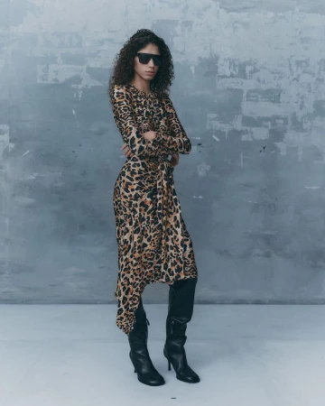 Woman with curly hair wearing sunglasses and a leopard print dress, standing indoors against a textured grey wall in a fashion pose.