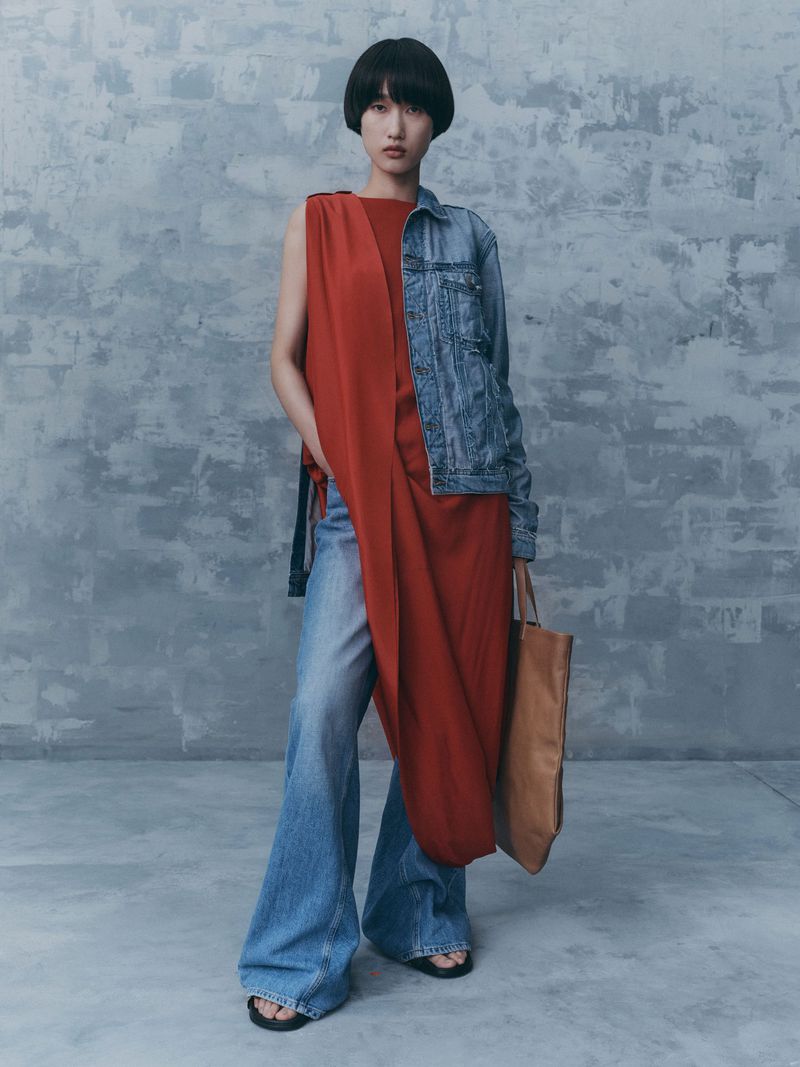 A brunette woman in a red dress, blue jean jacket, and jeans stands against a gray background with a brown tote bag.