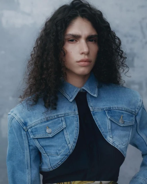 Portrait of a curly-haired person wearing a light-blue denim jacket, posing against a textured gray background, in a fashion editorial style.