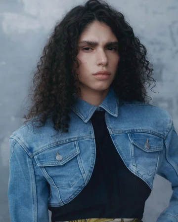 Portrait of a curly-haired person wearing a light-blue denim jacket, posing against a textured gray background, in a fashion editorial style.