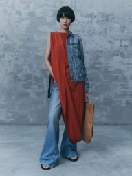 Model with dark bob hairstyle wearing a red dress, denim jacket and flare jeans holding a tan tote bag against a grey textured wall.