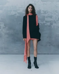 Model with curly dark hair wearing a black sleeveless top, pink scarf, and black boots against a textured grey wall