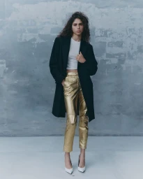Model with long curly hair wearing a black coat, white top, and metallic gold trousers against a textured grey wall