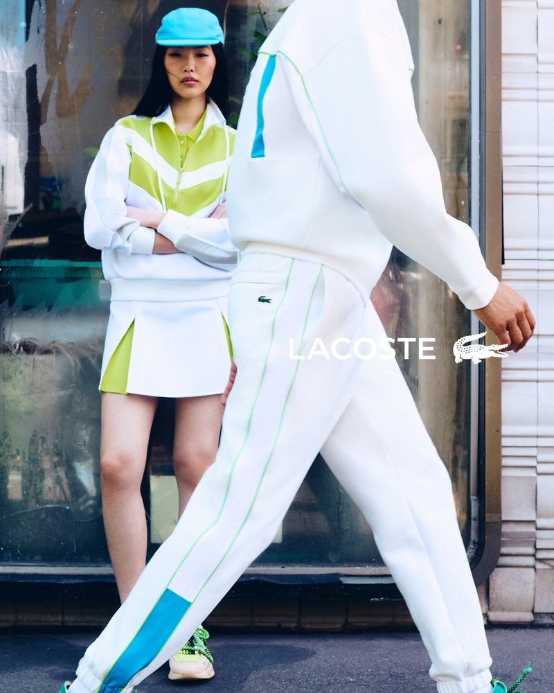 Two models in Lacoste sportswear in an urban setting, one wearing a light blue cap, skirt, and jacket, the other in white sweatpants and sweatshirt.