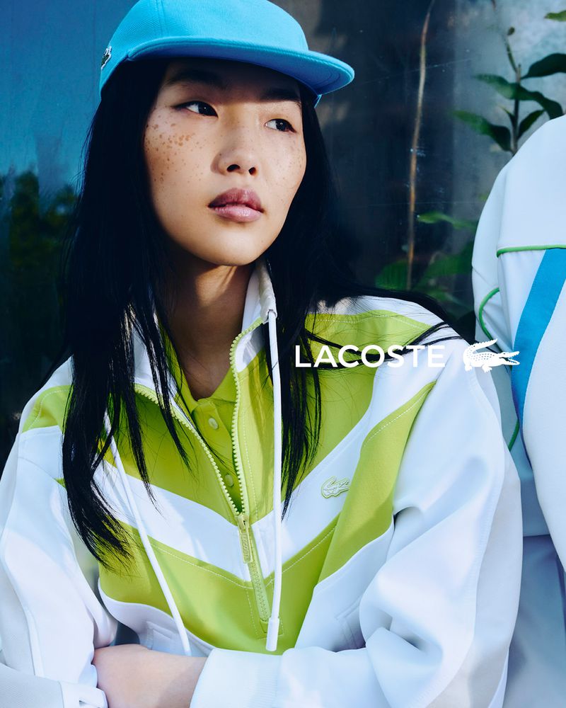 Close-up of Asian model wearing a turquoise baseball cap and white, green, and yellow Lacoste jacket against a blurred background.