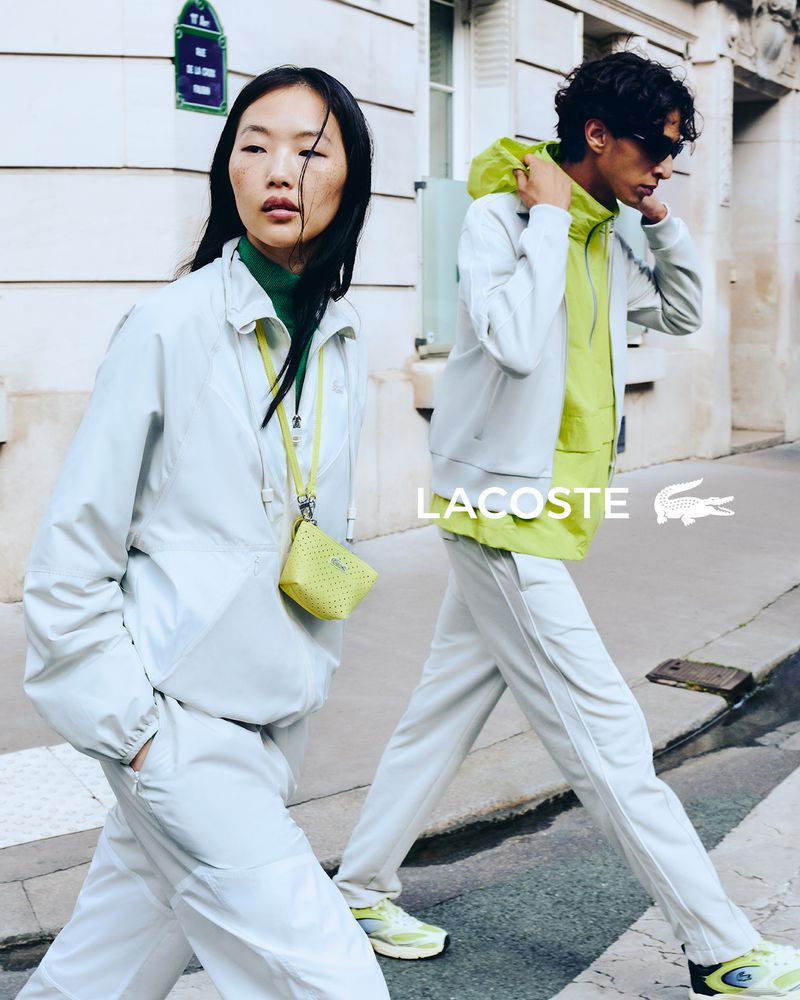 Two adult models in athletic wear walk on a street. One wears a light grey jacket and pants, the other a light grey jacket and pants with a yellow hoodie.