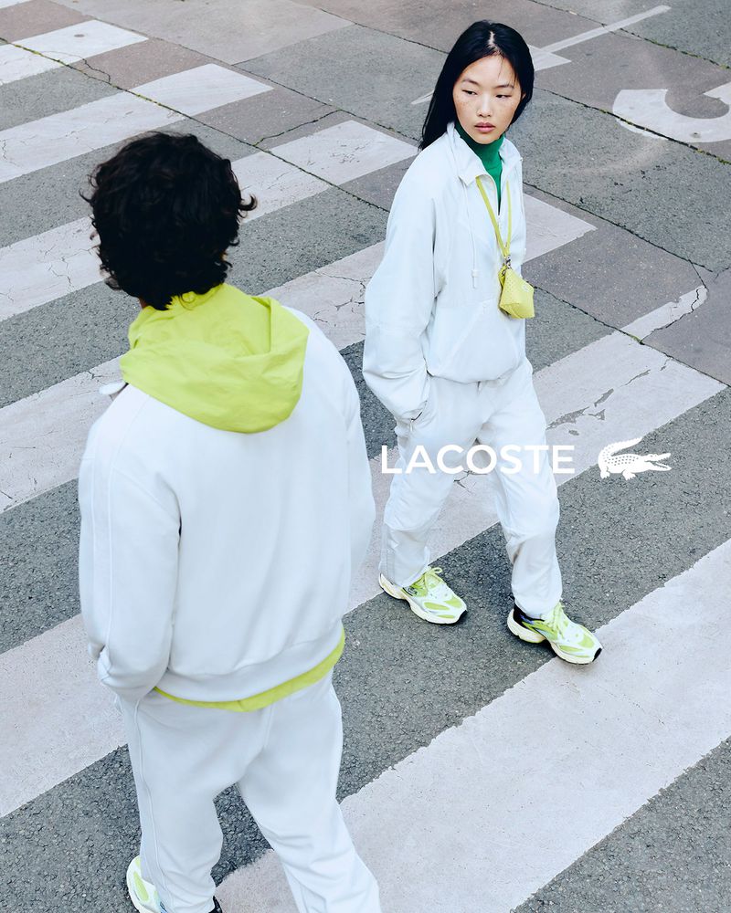 Two young adults in light colored activewear walking across a crosswalk in a street with a Lacoste logo overlayed on the image.