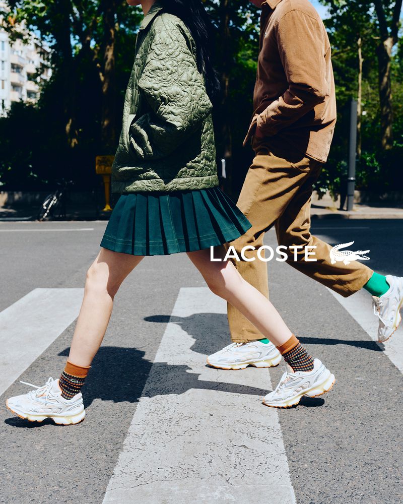 Two models walking across a street crosswalk wearing Lacoste apparel including a green quilted jacket and pleated skirt