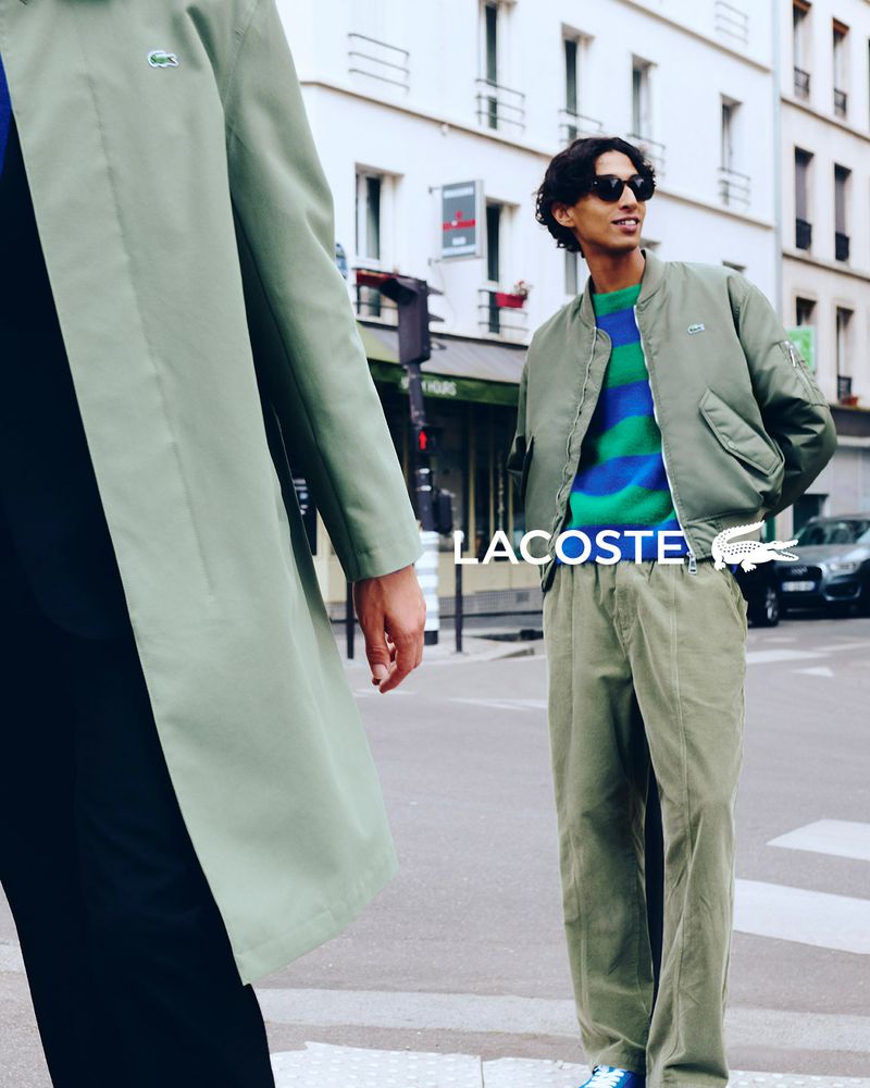 Two people wearing Lacoste apparel on a city street. One person wears a sage green trench coat, and the other wears a bomber jacket.
