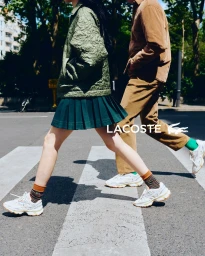 Two models walking across a street crosswalk wearing Lacoste apparel including a green quilted jacket and pleated skirt