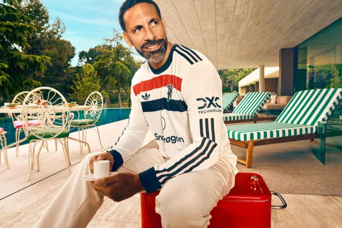 Rio Ferdinand lounges outside, holding a coffee cup, dressed in an Adidas long-sleeve shirt and off-white pants, on a sunny day.
