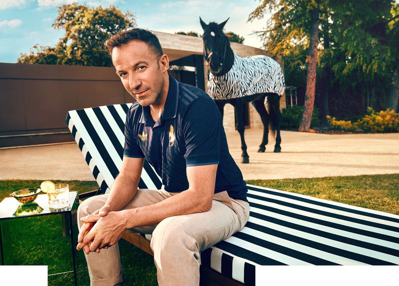 An adult male sitting on a striped lounge chair in an outdoor setting with a horse wearing a zebra-print blanket in the background.