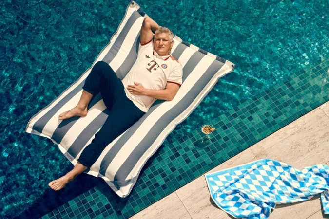 Adult male relaxes on striped inflatable raft in outdoor pool with turquoise water, glass of cocktail, and blue-white diamond-patterned towel.