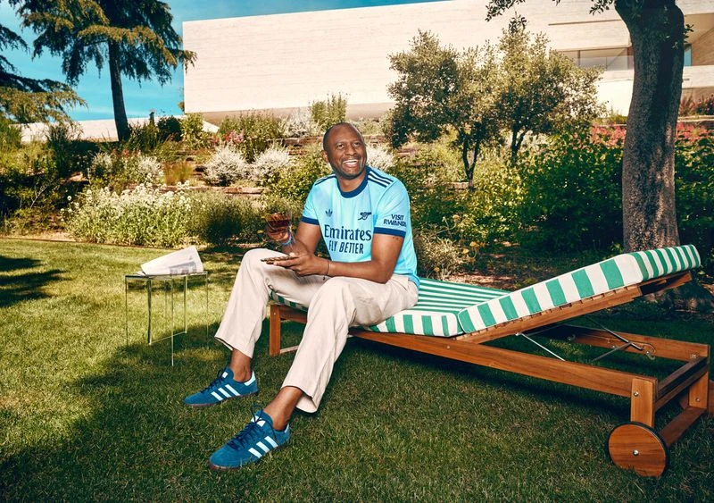 Adult male poses in a garden in an athletic shirt and khaki pants while sitting on a striped lounge chair with a drink and phone.
