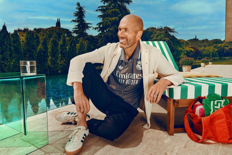Man in soccer jersey posing outdoors in casual wear, smiling and seated with trees and pool in the background on a sunny day.