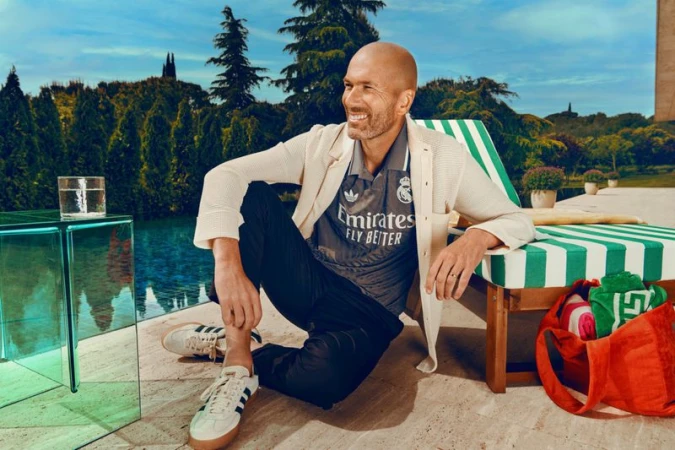 Man in soccer jersey posing outdoors in casual wear, smiling and seated with trees and pool in the background on a sunny day.