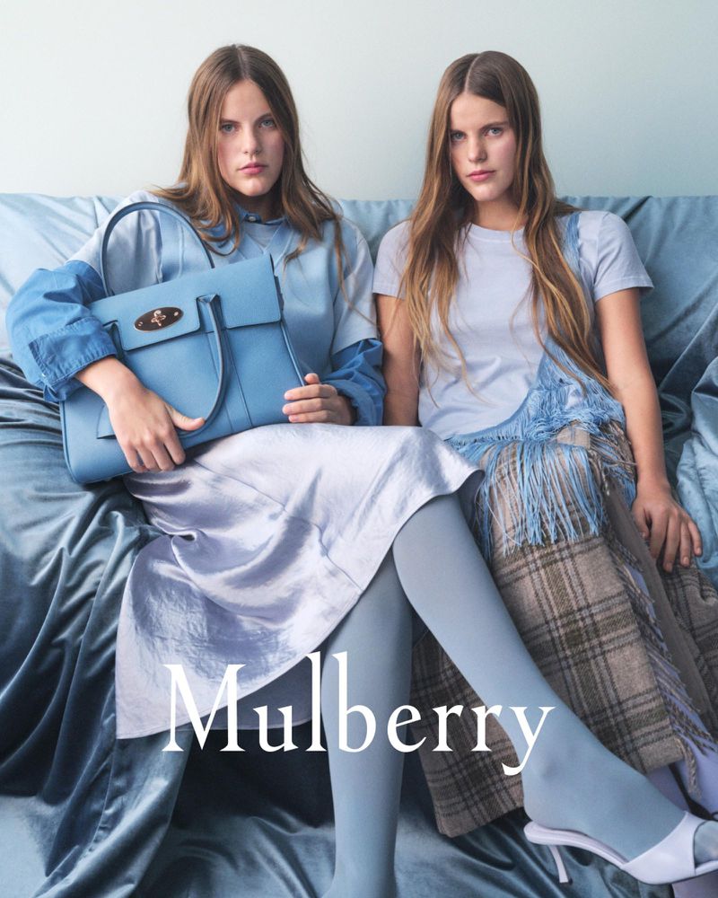 Two adult women model a blue leather satchel-style Mulberry handbag on a blue velvet couch. Fashion advertisement.
