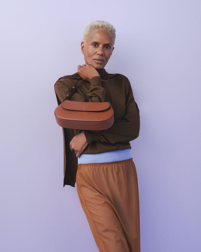A woman with short blonde hair poses with a brown leather purse against a lilac background, wearing a brown sweater and pants.