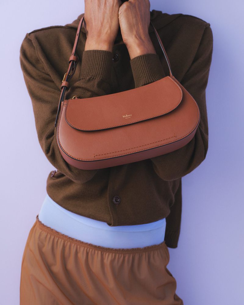 Person presents a brown Mulberry handbag, showcasing its design and color against a light purple background with warm studio lighting.