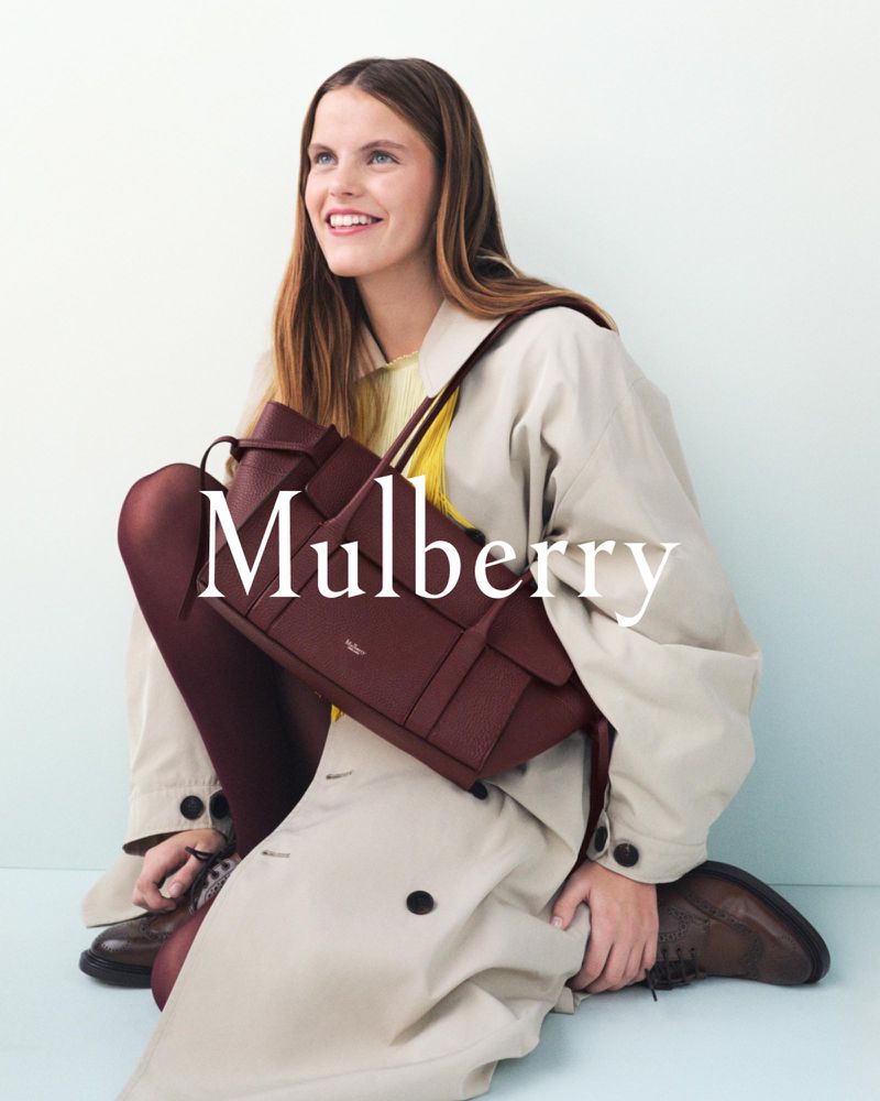 Smiling woman poses with a Mulberry handbag, wearing a tan trench coat, brown tights, and shoes against a light blue backdrop.