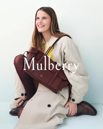 Smiling woman poses with a Mulberry handbag, wearing a tan trench coat, brown tights, and shoes against a light blue backdrop.
