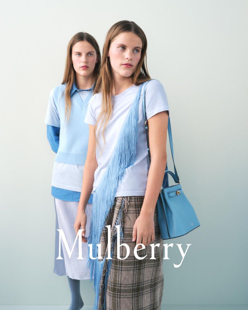 Two female models posing for the Mulberry T2 campaign against a pale gradient backdrop wearing blue apparel and holding a blue handbag